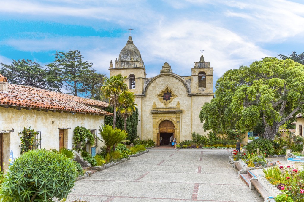 Facade of an adobe church with a stone courtyard and gardens.