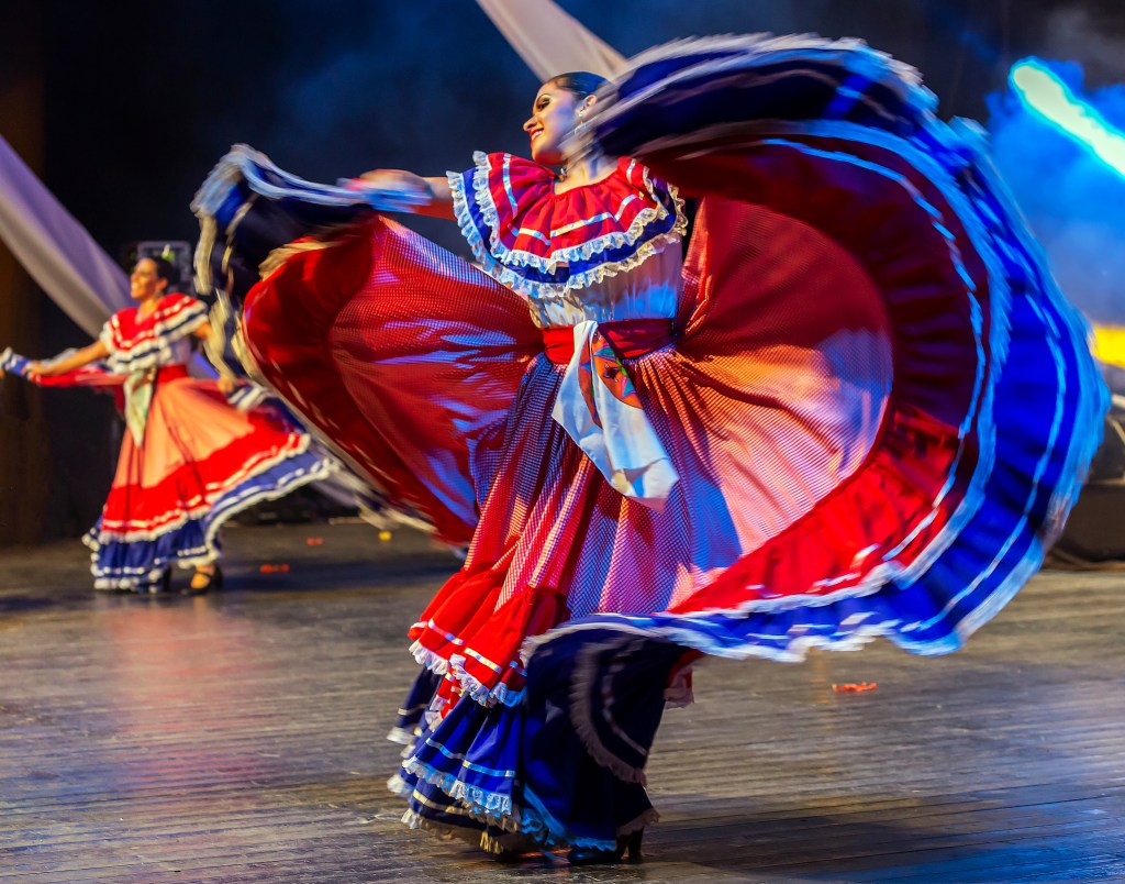 a performer in traditional costa rican skirts flares her arms mid-dance on a stage, with the red and blue fabric of her outfit swirling above her head
