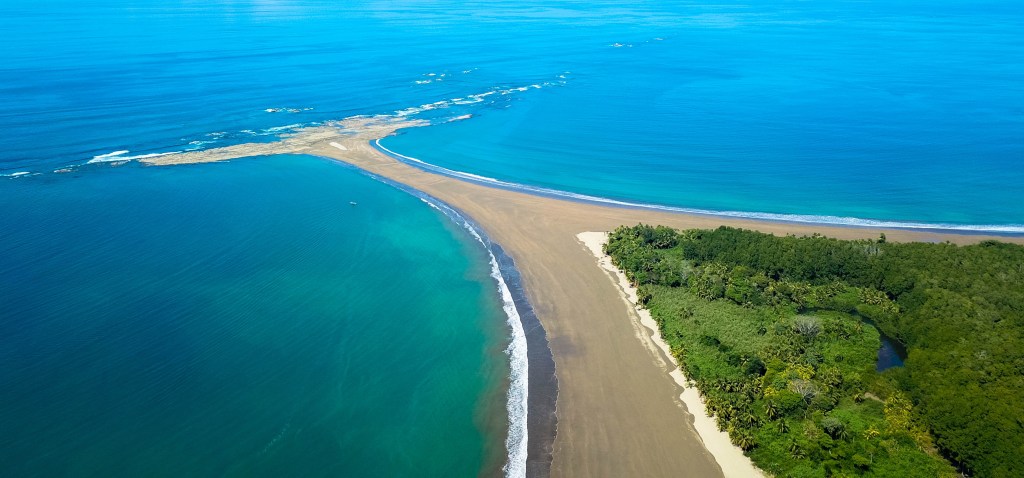 an arial view of a beach point that leads into a perpendicular sandbar - creating a peculiar whale tail shape. the sand is framed on both sides by deep blue open ocean