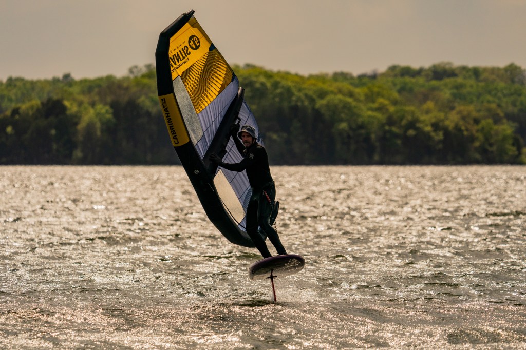 a wind surfer on the water on a windy day, with choppy water beneath and green trees in the background