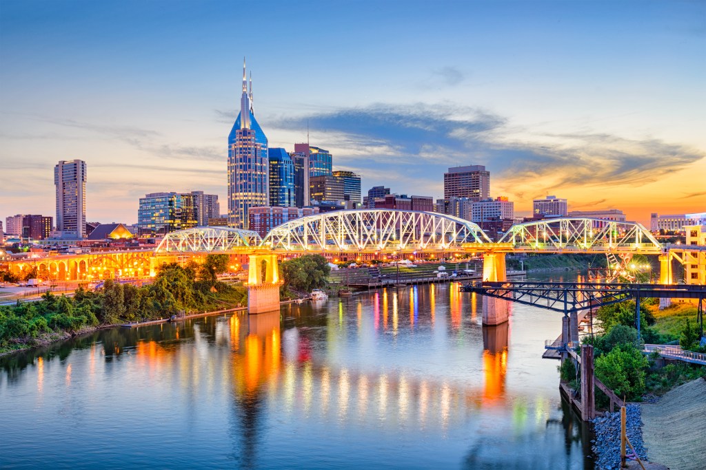 The Nashville skyline at sunset, with he warm orange lights of the John Seigenthaler Pedestrian Bridge in the foreground, reflecting off the surface of the Cumberland River.