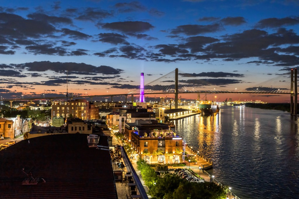 a view of the Savannah skyline from a roof deck after the sun has just set, with dark clouds still visible against the evening sky and the city streets lit up invitingly below