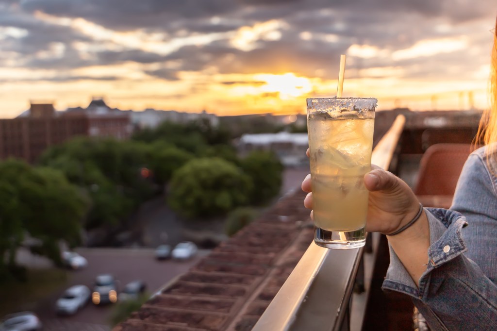 A hand holding a yellow alcoholic beverage with a straw and salted rim at sunset at a rooftop bar, with the tree-lined road beneath out of focus