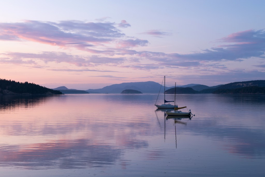 the sunsetting purple and pink of the calm waters of the San Juan islands, with two sailboats floating in the foreground and misty islands in the far horizon.