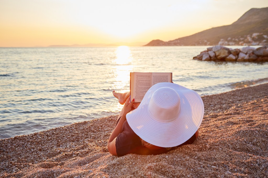 a woman in a big white sun hat reads a book while relaxing on the beach, overlooking a sunset over the rocky coastline