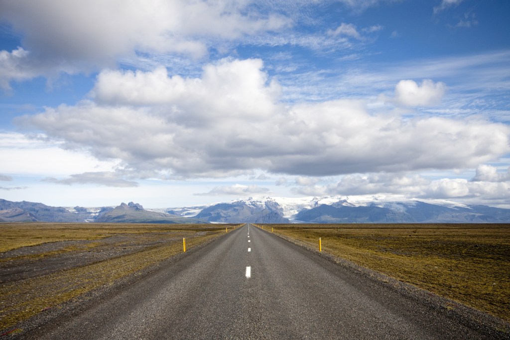 A two-lane road leading between flat brown fields to snowy mountains in the distance under blue sky with puffy white clouds