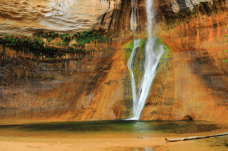 Image of skinny waterfall flowing down green and orange rock walls into a pool.