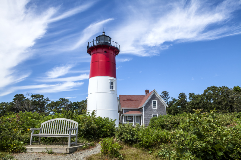 A red and white lighthouse next to a small grey-shingled house surrounded by gardens and a wooden bench under blue sky with wispy white clouds.