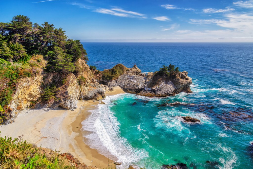 A sandy cove and turquoise ocean bordered by rocky cliffs covered in trees