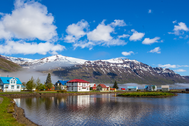 Picturesque small town of white buildings with colorful roofs along body of water with snowcapped mountains in the distance.